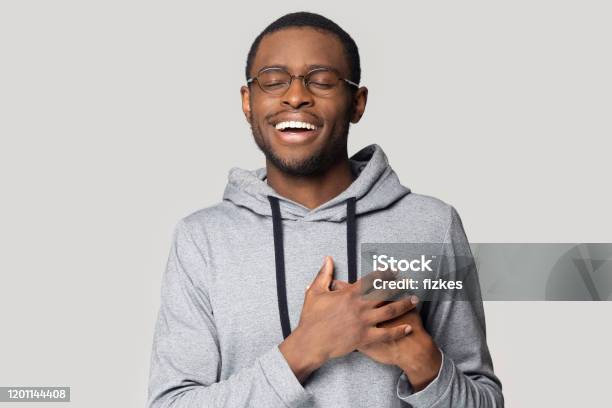 Happy african American man in glasses isolated on grey studio background keep hands at heart chest feel grateful, thankful black male in spectacles stand with eyes closed smiling thanking to god